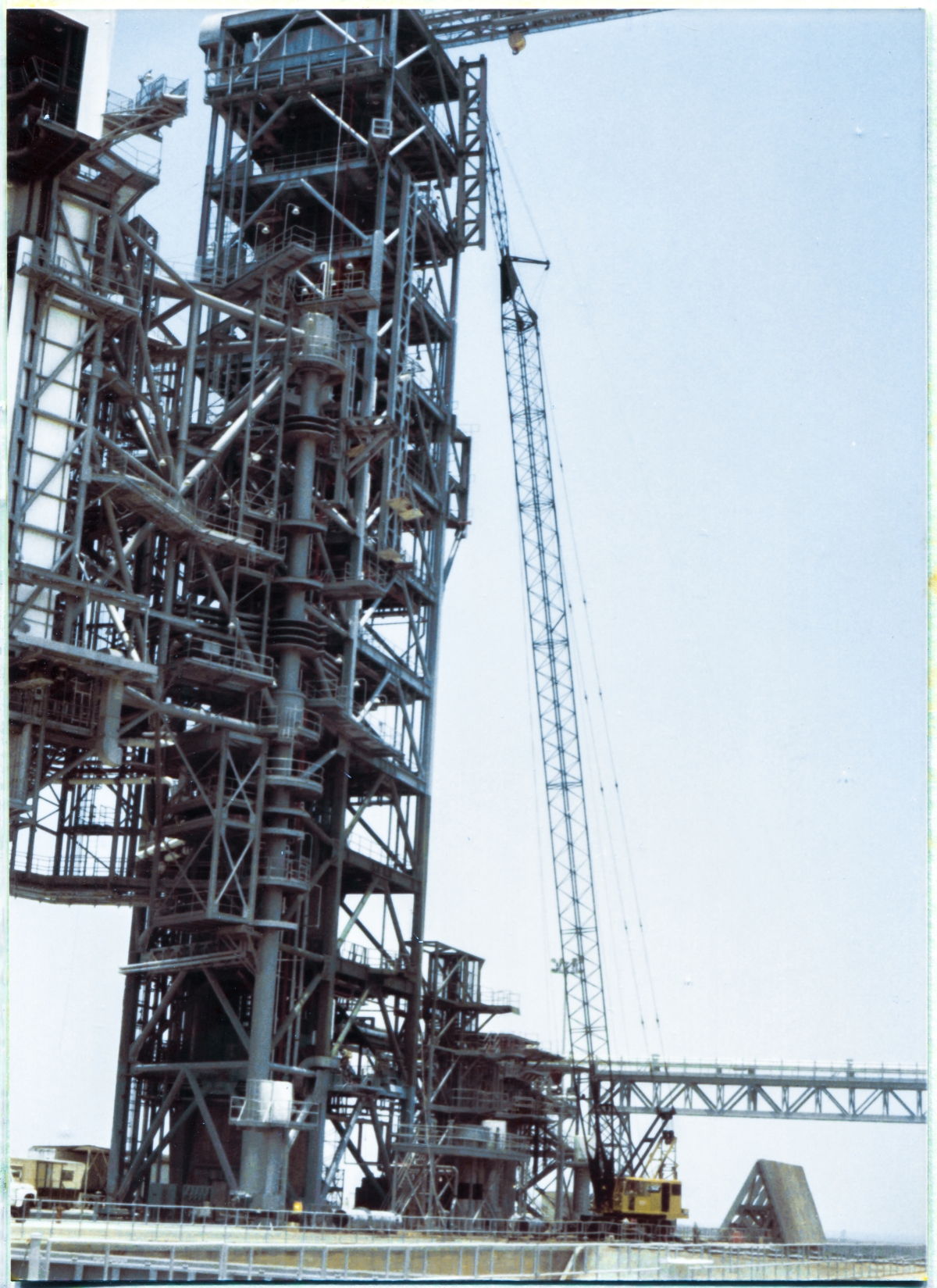 Image 076. The GOX Arm Strongback has reached its final elevation at the top of the Fixed Service Structure at Space Shuttle Launch Complex 39-B, Kennedy Space Center, Florida, with the tops of its twin W24x104 longitudinal members located just above elevation 300'-0”, which is the level of the deckplates on top of the FSS. The tag line which we saw earlier, attached to its lower right corner, was walked by the irownworker holding on to it, down on the ground, over to a position where it became draped across the front of the FSS, in direct contact with it, allowing other ironworkers up on the tower to reach it from up there, gather it in and get it out of the way, and continue using it to keep pulling the Strongback around, more or less to an orientation where you see it in this photograph. From there on, the tag line becomes unnecessary, is removed, and everything from here on is done with come-alongs, bringing much greater force to bear, getting the Strongback into its final position for welding and bolting it to the tower. As background, the colossal size of the FSS, spilling out of the frame up above, becomes apparent, and our once quite-large Strongback has now been dwarfed into insignificance by it. Photo by James MacLaren.